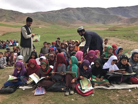 2 men handing out books to Afghan girls seated outside